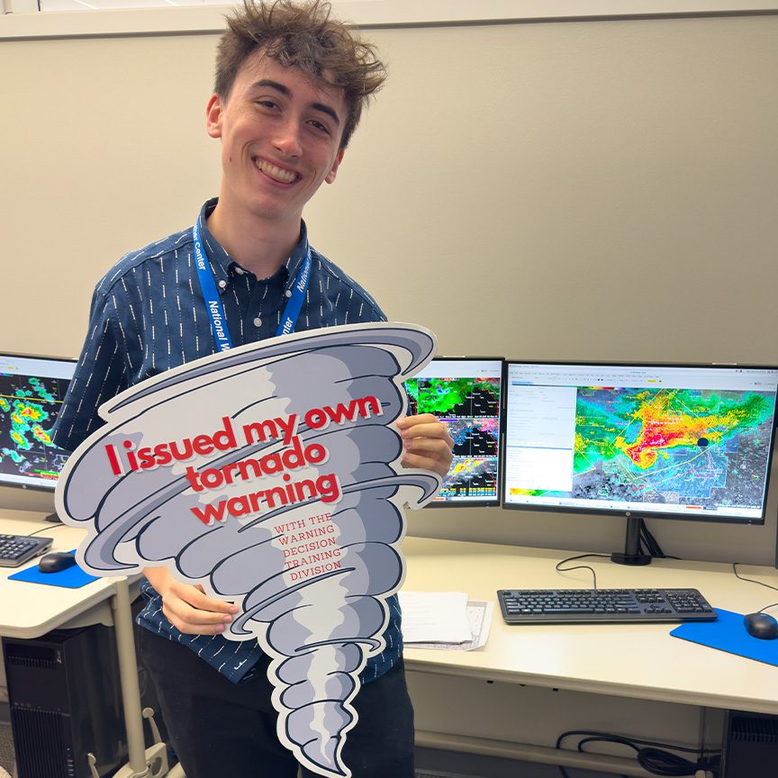 Ian Shank holding a sign in the shape of a tornado that reads "I Issued My First Tornado Warning"