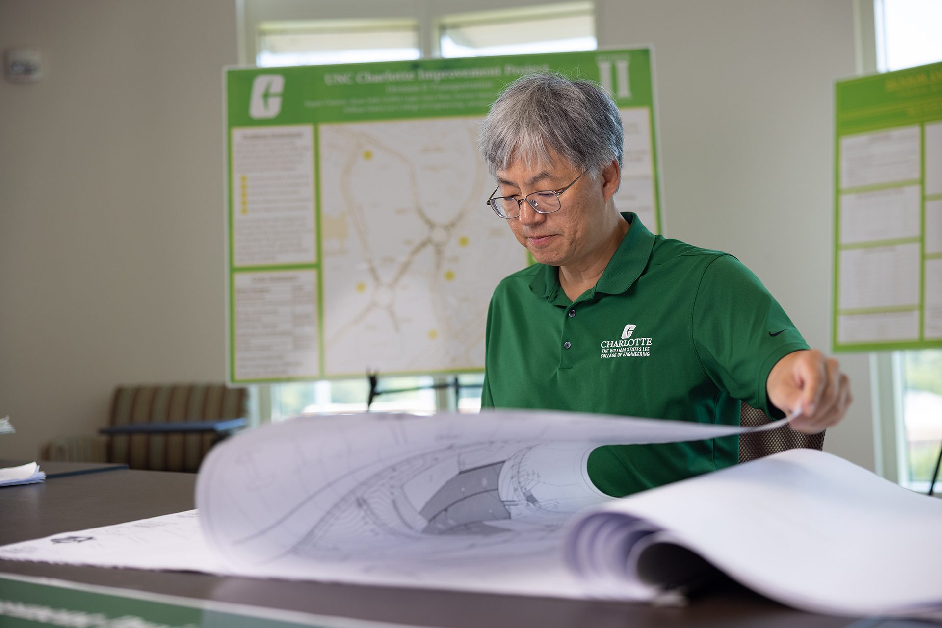 A man in a green UNC Charlotte College of Engineering shirt reviews large rolled blueprints at a table, with campus improvement project posters displayed behind him. {{brizy_dc_image_alt imageSrc=