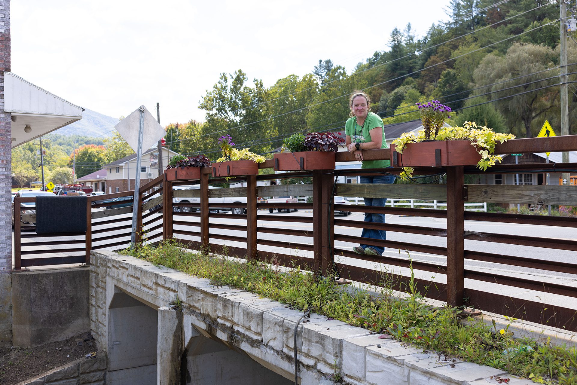 A woman in a green shirt leans on a wooden railing decorated with planter boxes full of colorful flowers, standing above a stone drainage channel beside a road in a small town. {{brizy_dc_image_alt imageSrc=