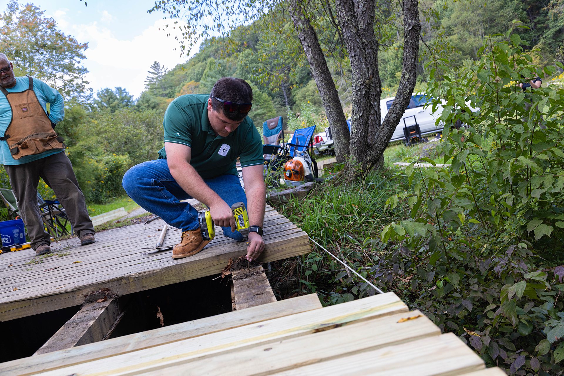 A man in safety glasses and a green shirt crouches on a wooden deck, using a power drill to repair boards, while another man wearing an apron observes nearby. {{brizy_dc_image_alt imageSrc=