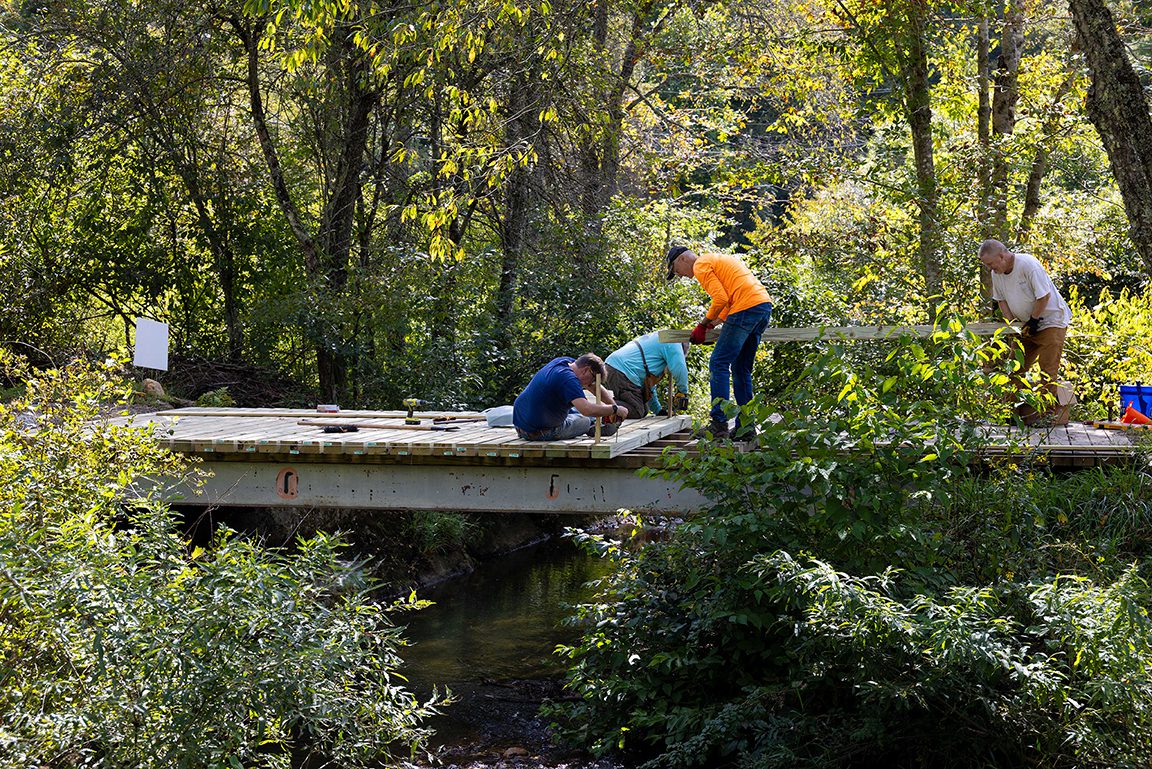 A group of four people works together repairing a wooden footbridge in a wooded area, hammering and lifting boards over a stream below. {{brizy_dc_image_alt imageSrc=