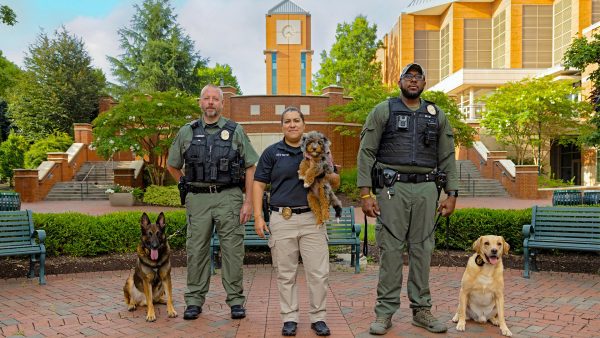 Three police officers pose together in front of a campus clock tower with their dogs, including a German Shepherd, a small curly-haired dog, and a Labrador Retriever.