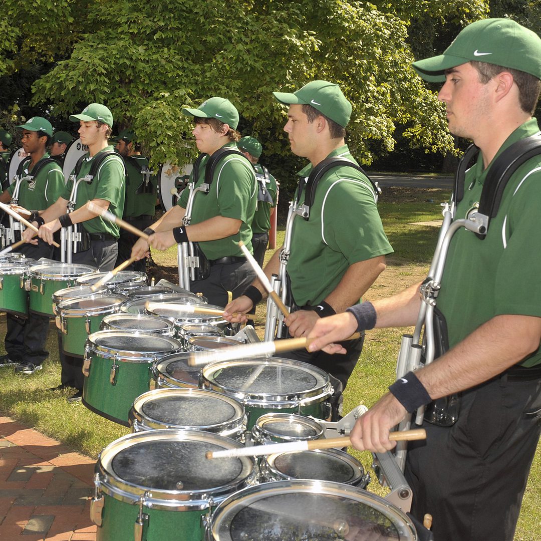 A row of UNC Charlotte drumline members performing outdoors in matching green polo shirts, black pants, and green baseball caps. Each is playing a set of tenor drums, with sunlight filtering through the surrounding trees.