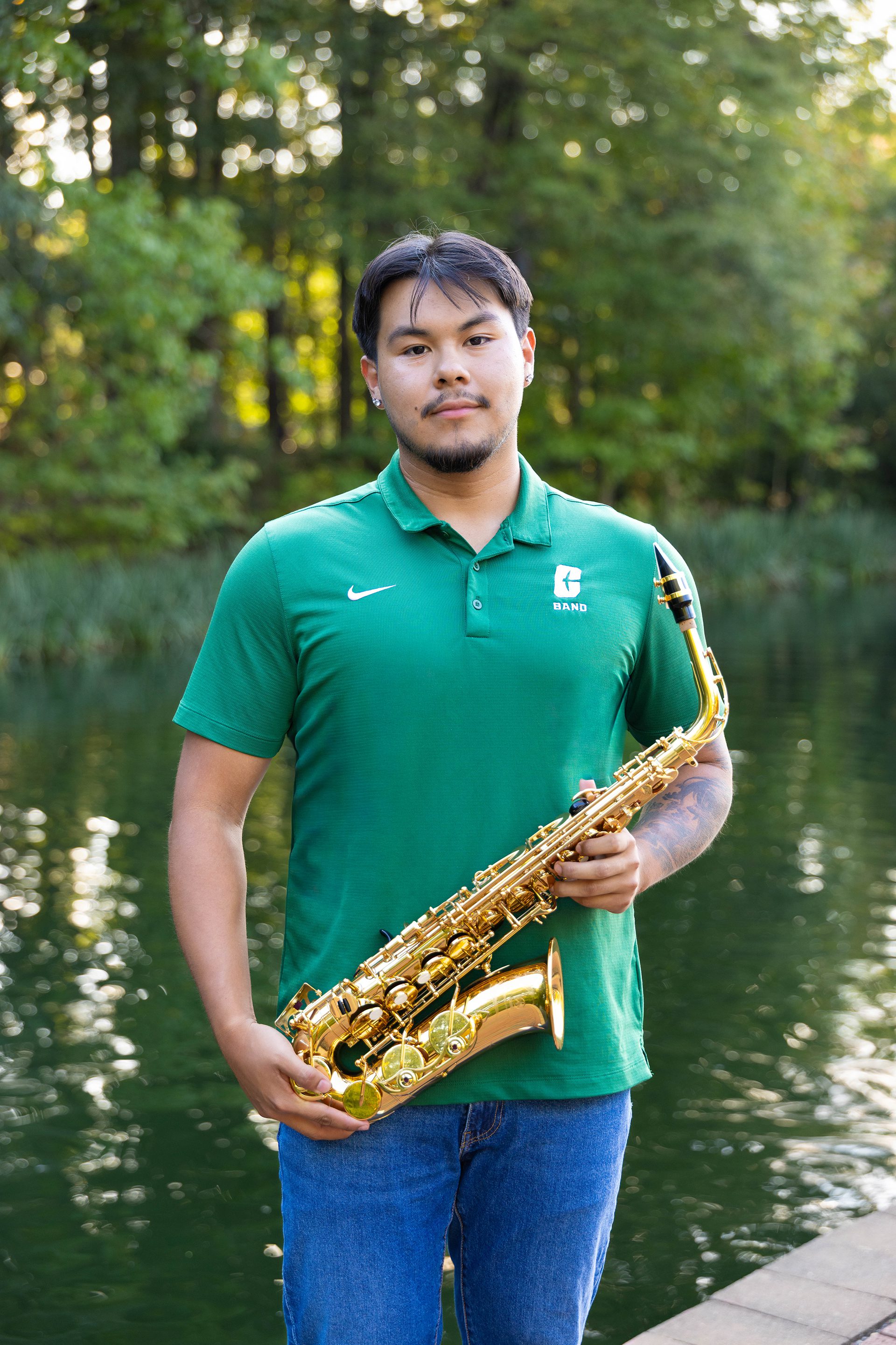 A UNC Charlotte student wearing a green “Band” polo stands by the campus lake, holding a golden alto saxophone with both hands and smiling slightly at the camera. Trees and water are visible in the background.