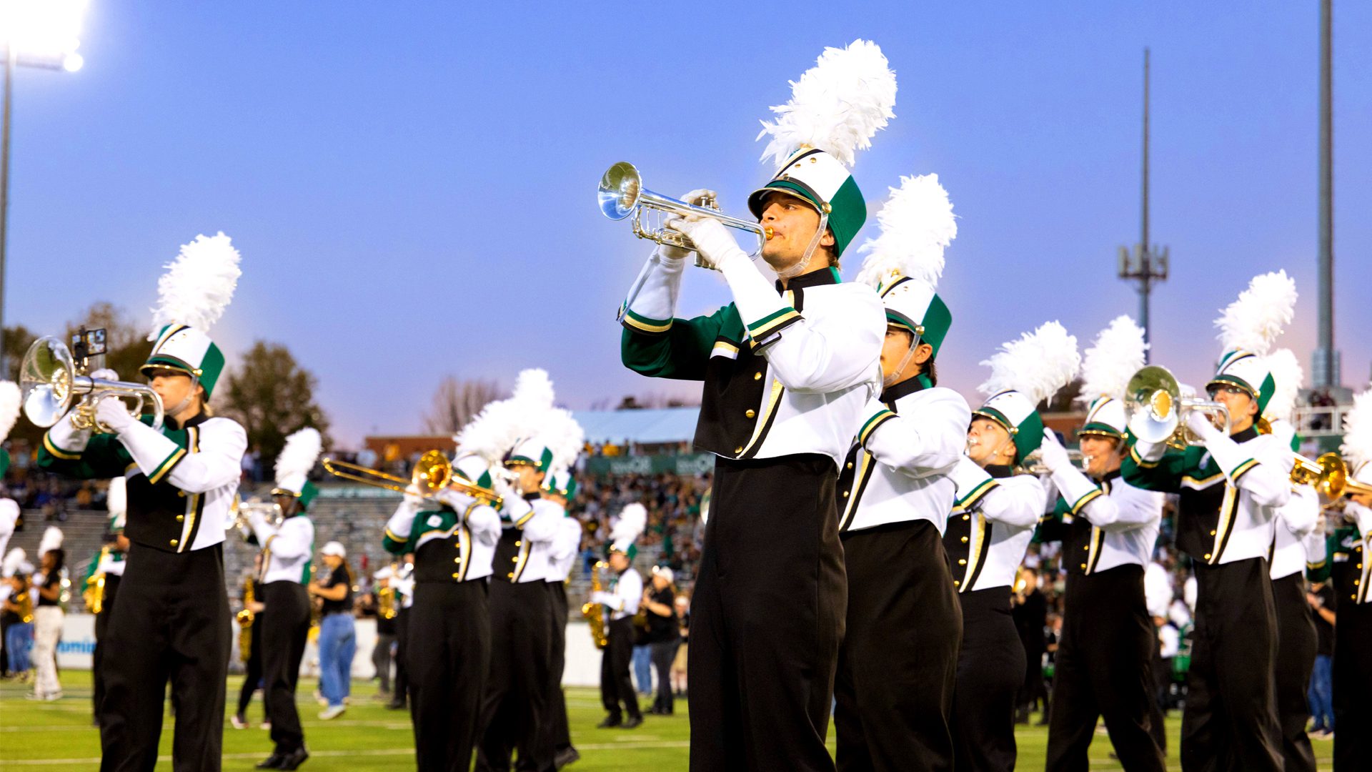 A university marching band performs on a football field during a game at sunset. Band members, wearing green, white, and black uniforms with tall white plume hats, play brass instruments in formation. The stadium lights shine brightly as a crowd watches from the stands in the background.