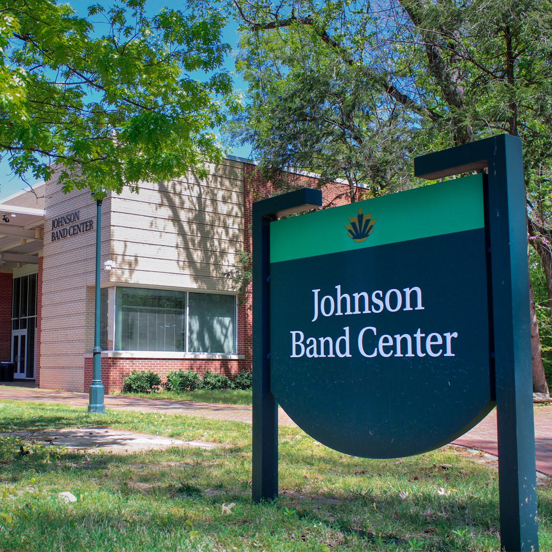 A sunny exterior view of the Johnson Band Center at UNC Charlotte, showing the modern brick and panel building behind a large green and black sign with the university's crown logo and the words “Johnson Band Center.”
