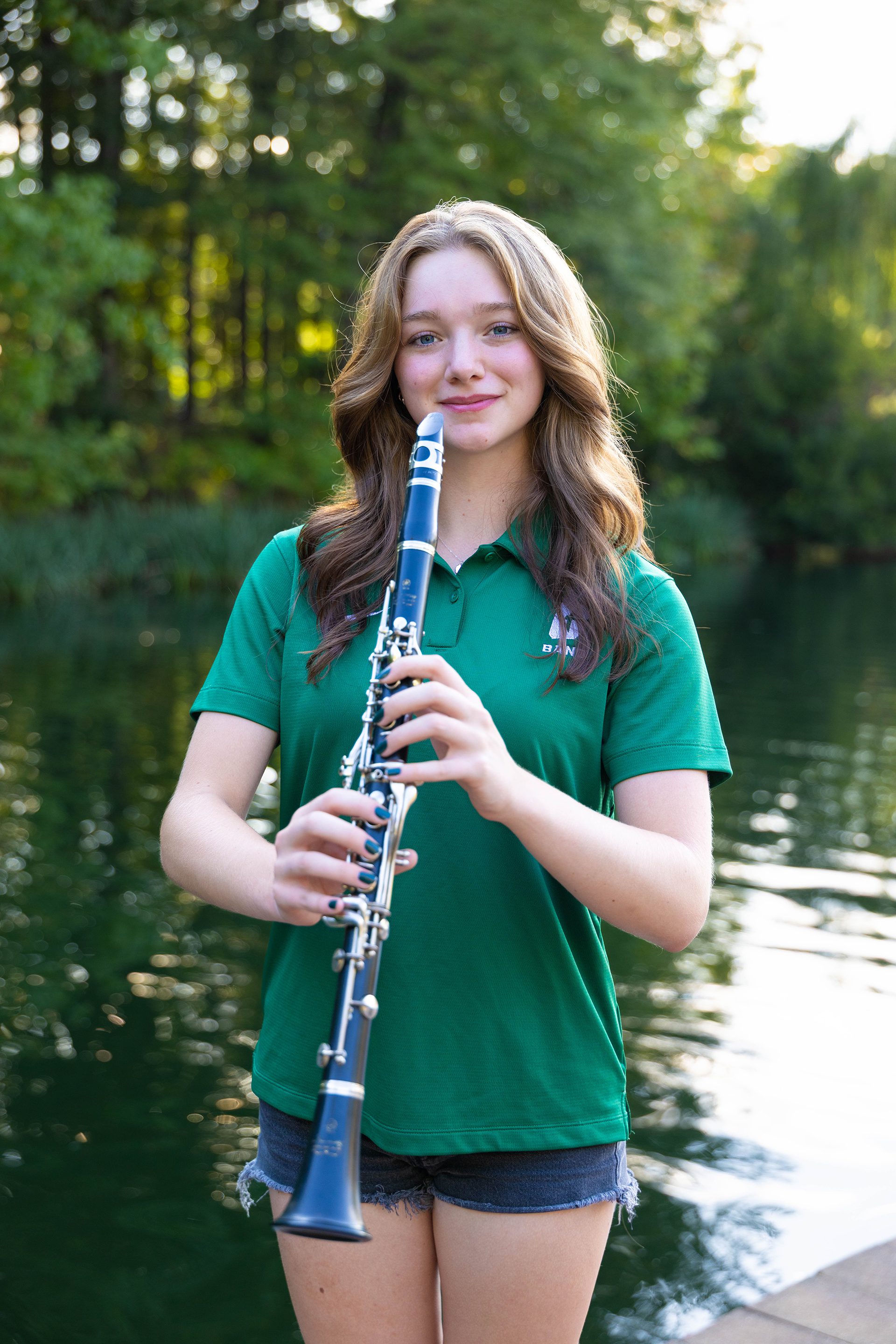 A student in a green UNC Charlotte “Band” polo and denim shorts holds a black clarinet with both hands, smiling softly while standing by the lake with trees in the background.
