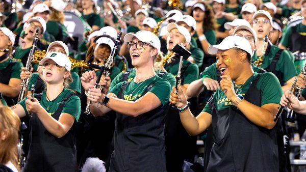 UNC Charlotte marching band members in green shirts, black overalls, and white hats perform enthusiastically in the stadium stands during a football game, smiling and playing their instruments with gold glitter on their faces.