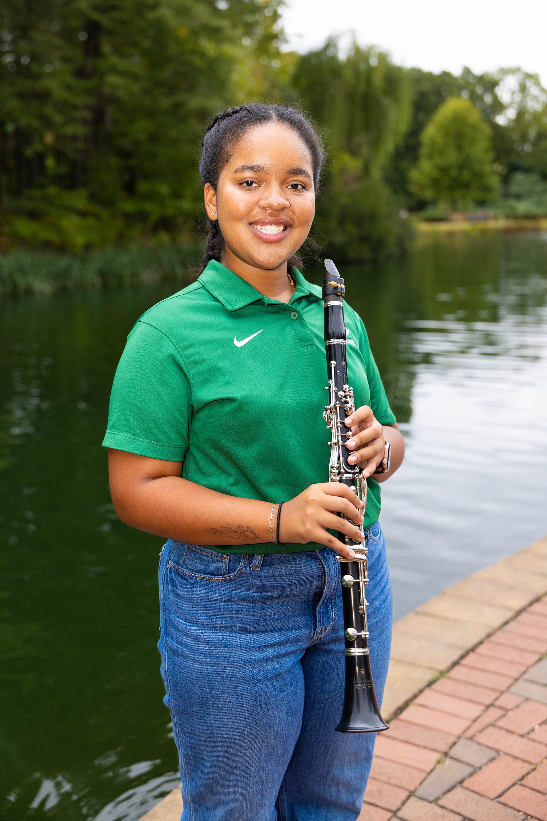 A student in a green UNC Charlotte polo and blue jeans stands on the lakefront path, smiling while holding a clarinet. The water and lush trees reflect a peaceful background.