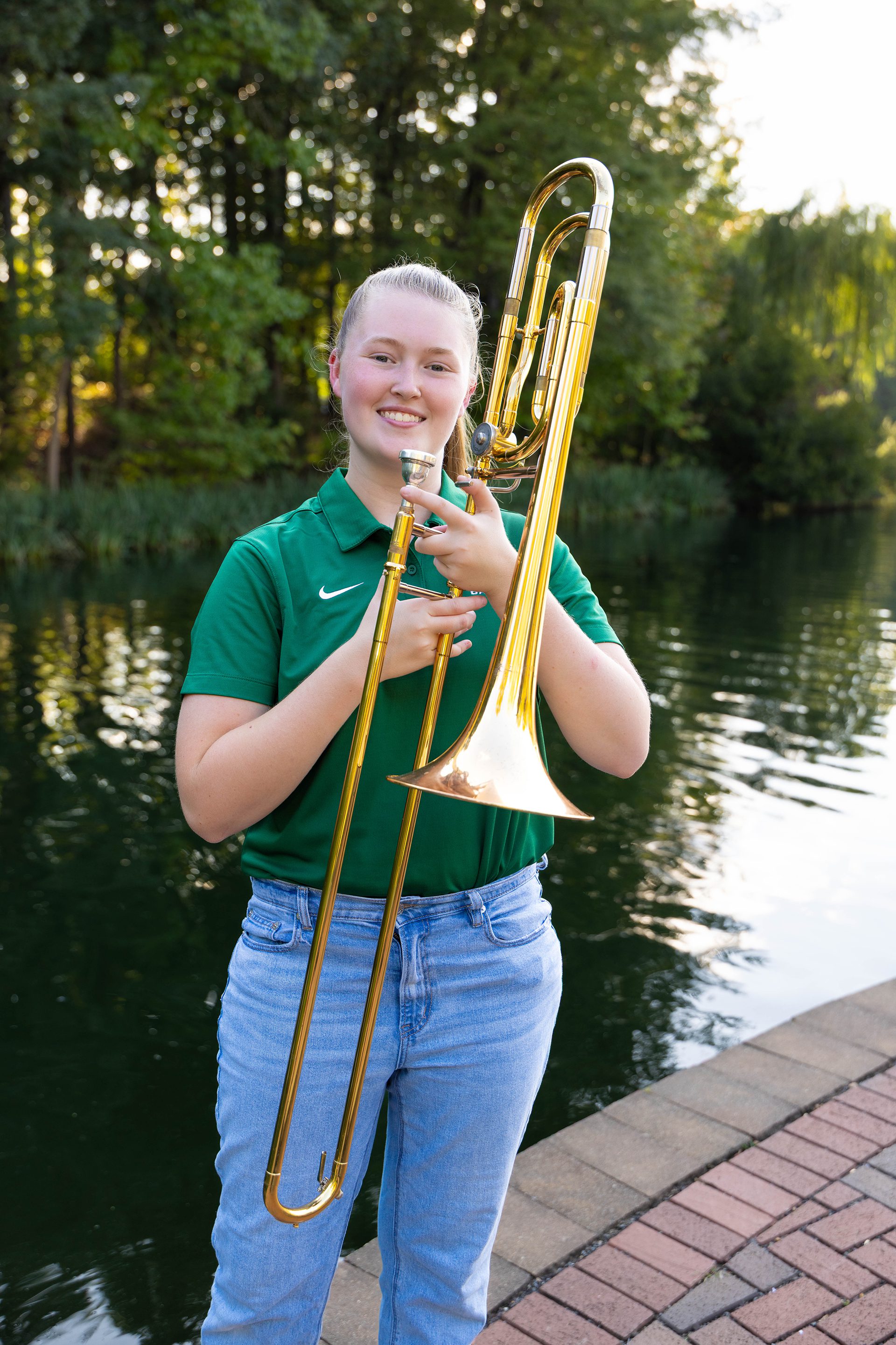 A UNC Charlotte band member in a green polo and light blue jeans smiles while holding a shiny brass trombone upright, standing on a brick path by the lake.