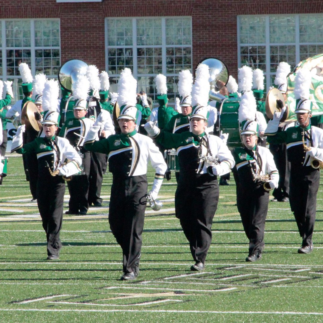 UNC Charlotte marching band members perform a halftime show on the football field in full green and white uniforms with tall white plume hats. The musicians play brass instruments while marching in formation in front of a red-brick building with large windows.