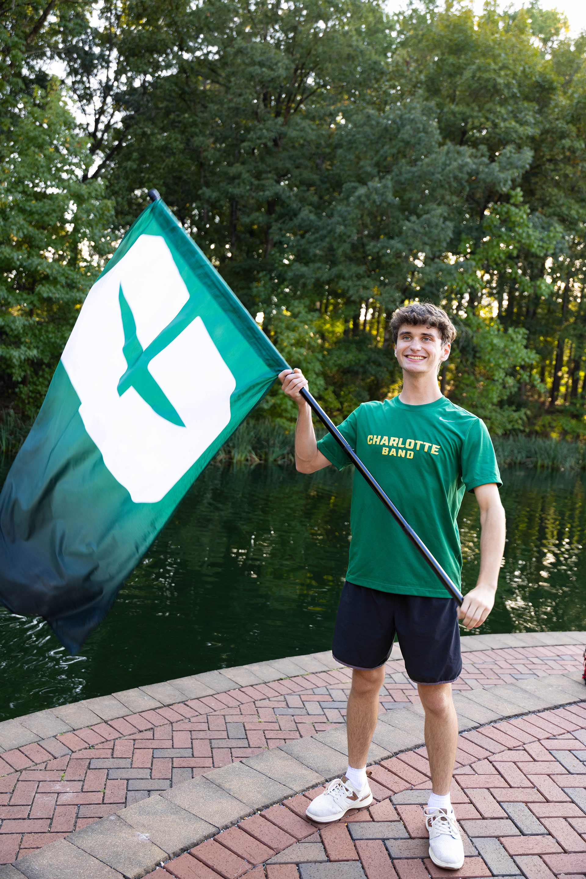 A student in a green “Charlotte Band” T-shirt and black shorts smiles while holding a large green and white UNC Charlotte flag, standing on the brick pathway by the campus lake.