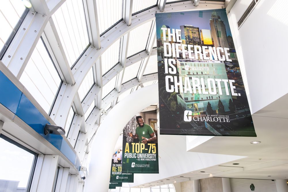Hanging banners in Charlotte Douglas International Airport featuring UNC Charlotte messaging, including “The Difference is Charlotte” and “A Top-75 Public University.”