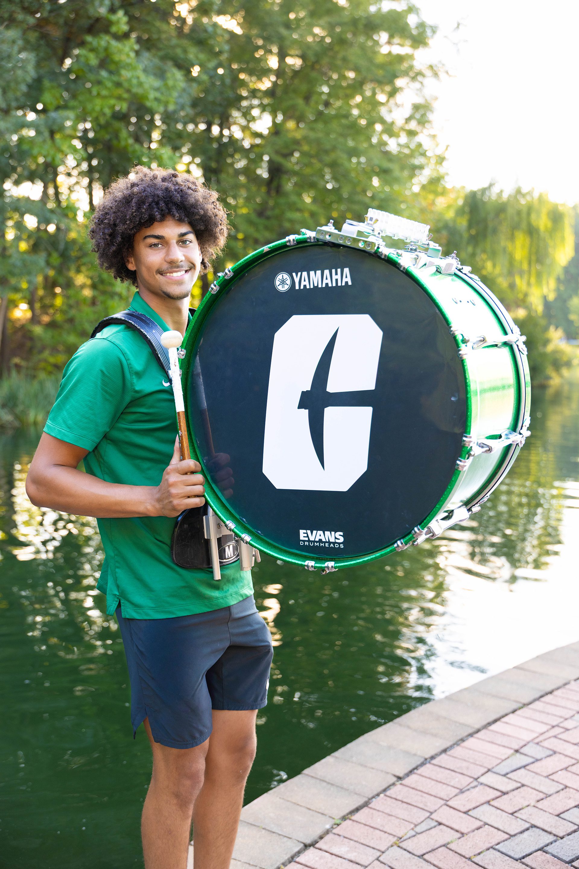 A student smiles while wearing a green UNC Charlotte polo and carrying a large bass drum emblazoned with the university’s athletics “C” logo. He stands beside the campus lake under green trees.