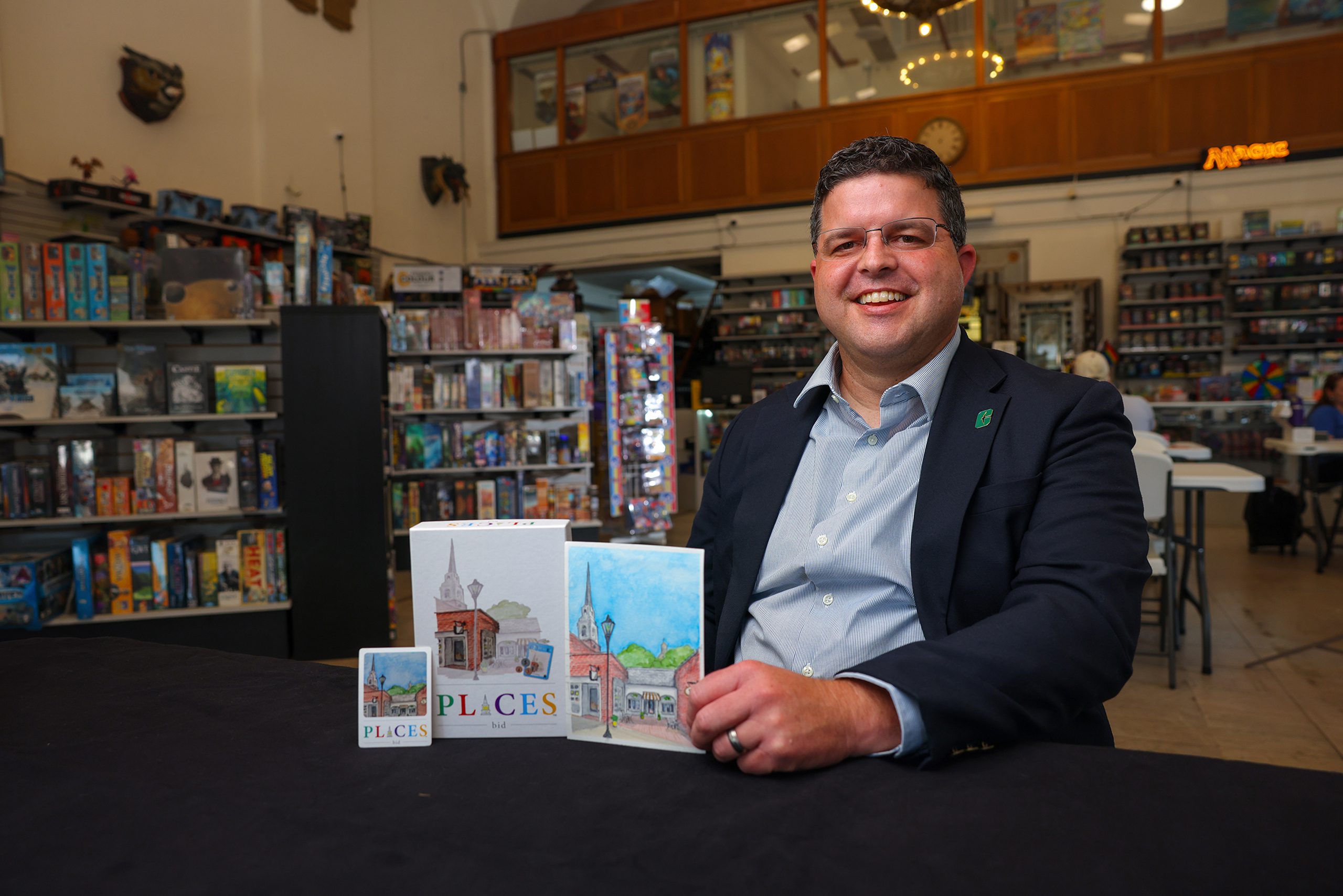 A smiling man in a UNC Charlotte lapel pin sits at a table in a board game store, showcasing a card game titled "PLACES" with illustrated packaging depicting a town square and church steeple.