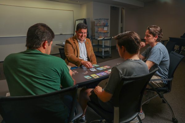 A group of four people sit around a table in a classroom, playing a colorful card game led by a man in a tan blazer wearing a UNC Charlotte lapel pin.