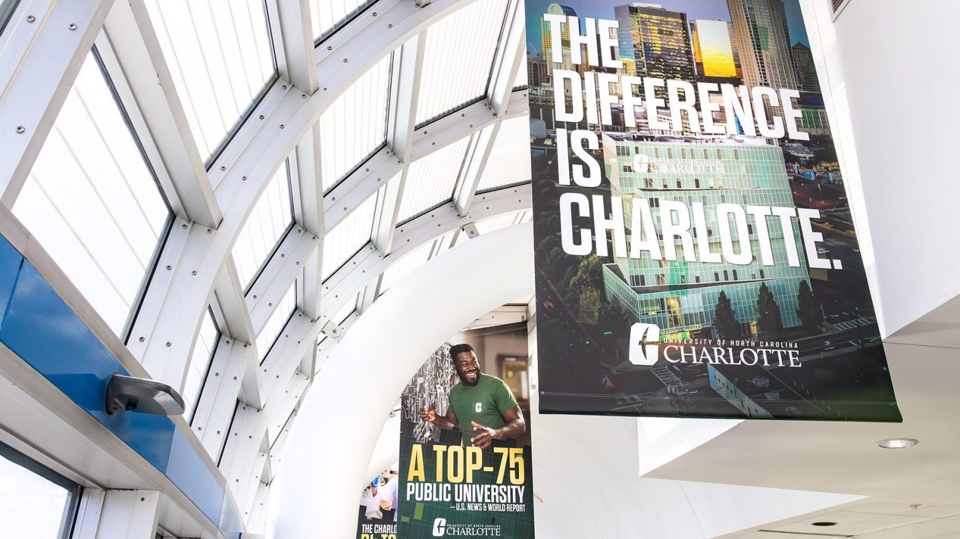 Hanging banners in Charlotte Douglas International Airport featuring UNC Charlotte messaging, including “The Difference is Charlotte” and “A Top-75 Public University.”