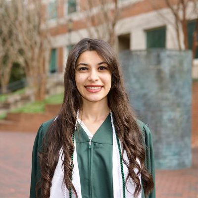 UNC Charlotte graduate wearing green regalia and cords, standing outside on campus with a smile.