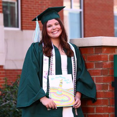 UNC Charlotte graduate in cap and gown holding a decorated graduation cap featuring “Oh, the Places You’ll Go!”