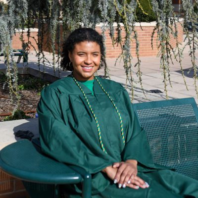 Smiling UNC Charlotte graduate sits on a campus bench in green regalia, under drooping tree branches.