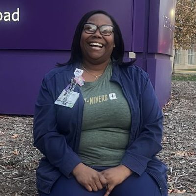 UNC Charlotte alumna wearing scrubs and a Niners shirt sits smiling outside near a purple building.