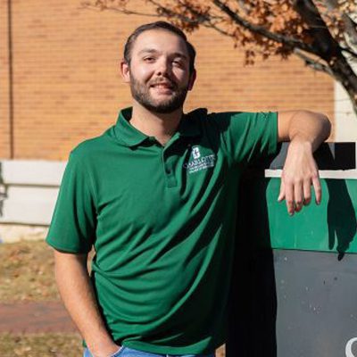 UNC Charlotte student in a green campus polo poses casually outdoors by a campus sign.