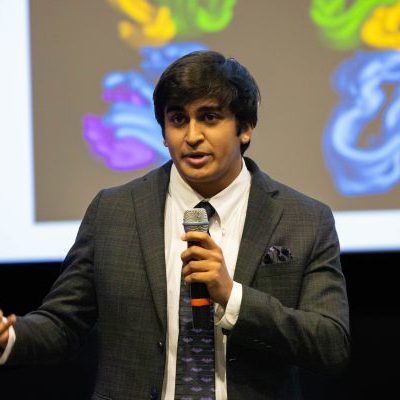 UNC Charlotte student in a suit speaks on stage with a microphone during a presentation with a colorful screen in the background.