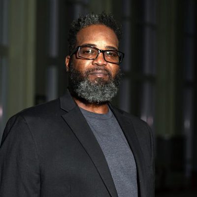 Portrait of a UNC Charlotte student wearing glasses and a blazer, standing in a dimly lit indoor space.