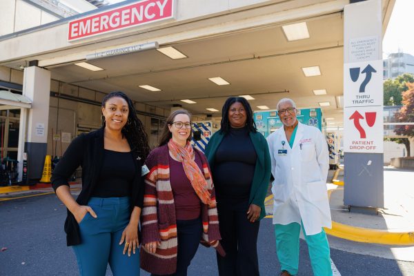 Four people, including a medical professional in a lab coat, stand smiling outside a hospital emergency entrance, with an ambulance visible in the background.