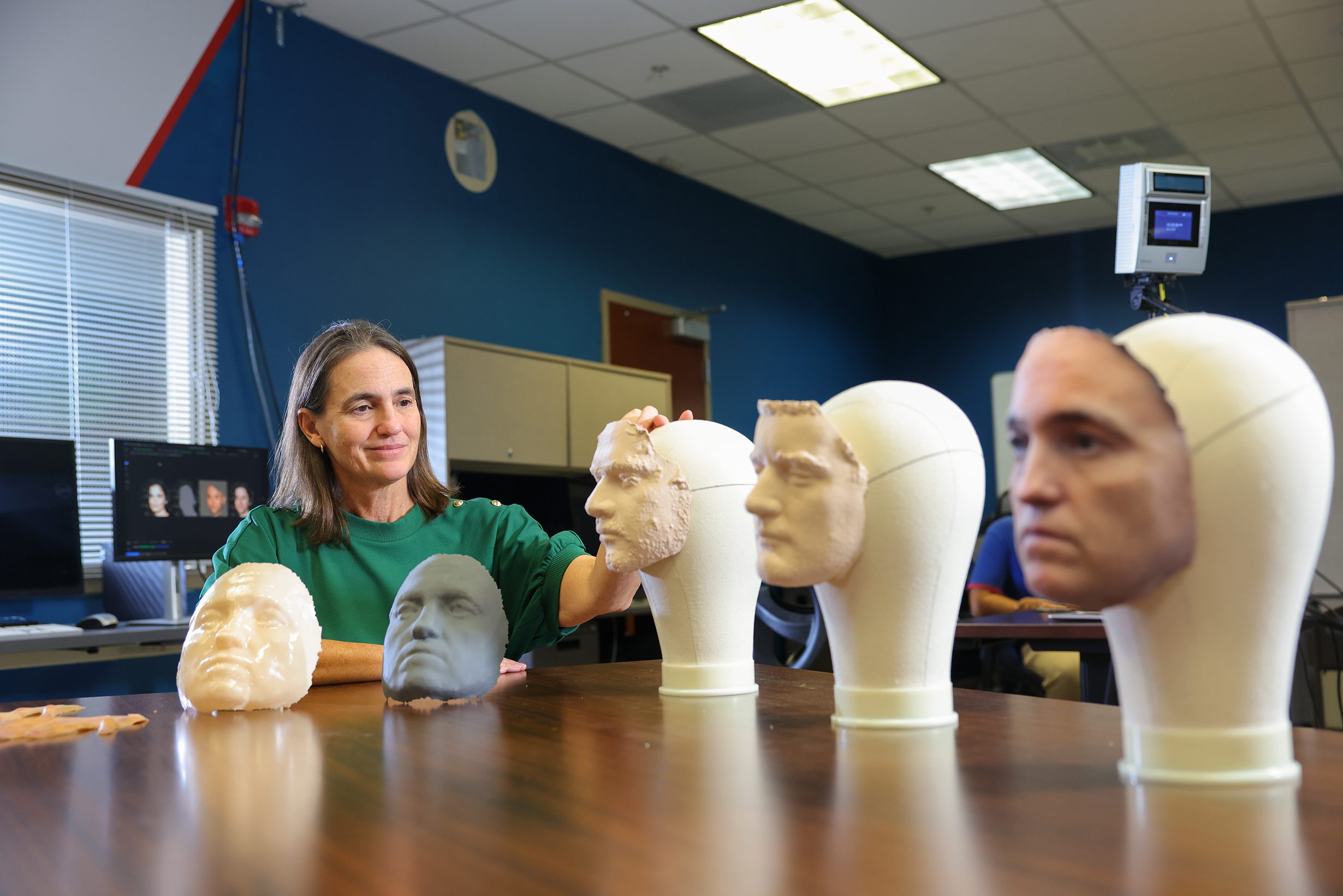 A woman in a green shirt examines a series of 3D-printed facial models mounted on mannequin heads in a research lab at UNC Charlotte, with monitors and lab equipment visible in the background.