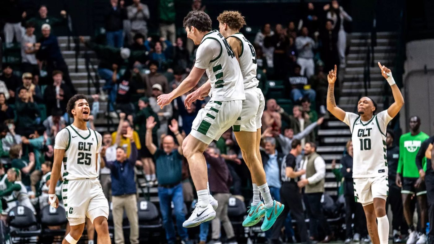Charlotte 49ers men’s basketball players celebrate during a game, jumping and cheering on the court while fans in the stands raise their arms in excitement behind them.