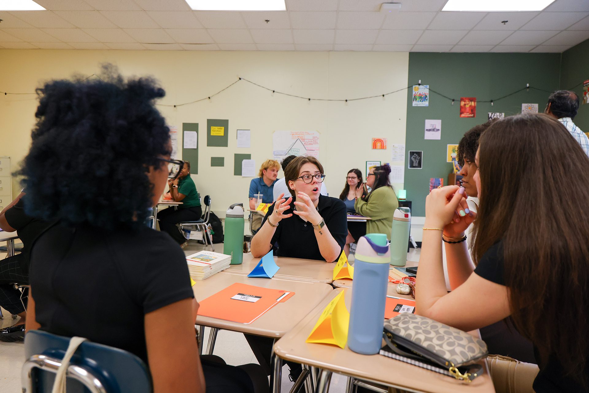 Instructor leading a discussion with students seated in small groups around desks in a classroom.