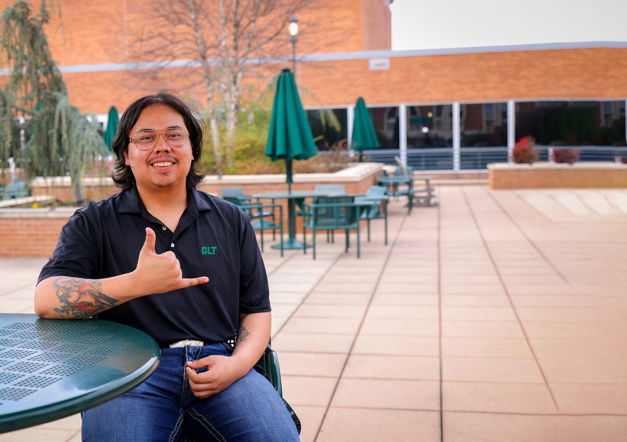 Transfer student sits at an outdoor campus table, smiling and making a hand gesture, with patio seating and campus buildings in the background.
