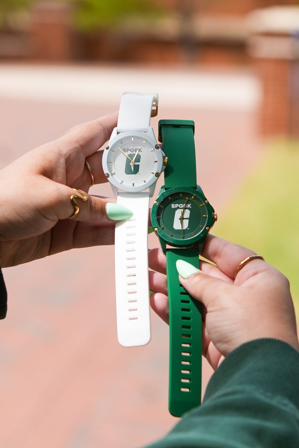 Hands holding two wristwatches — one white and one green — each featuring the UNC Charlotte “All-in-C” logo on the face.
