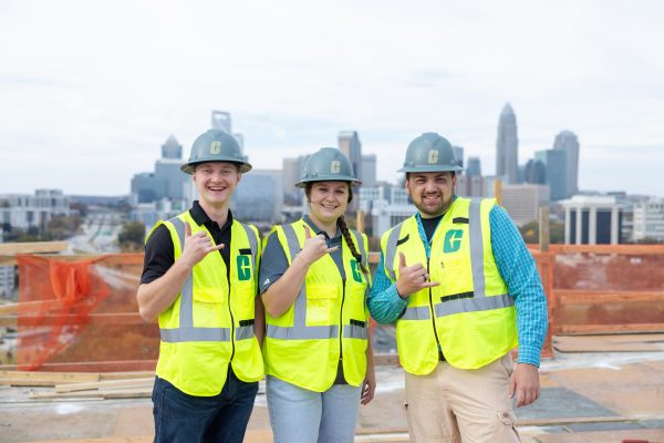 Three students in Charlotte hardhats stand in front of the city skyline.