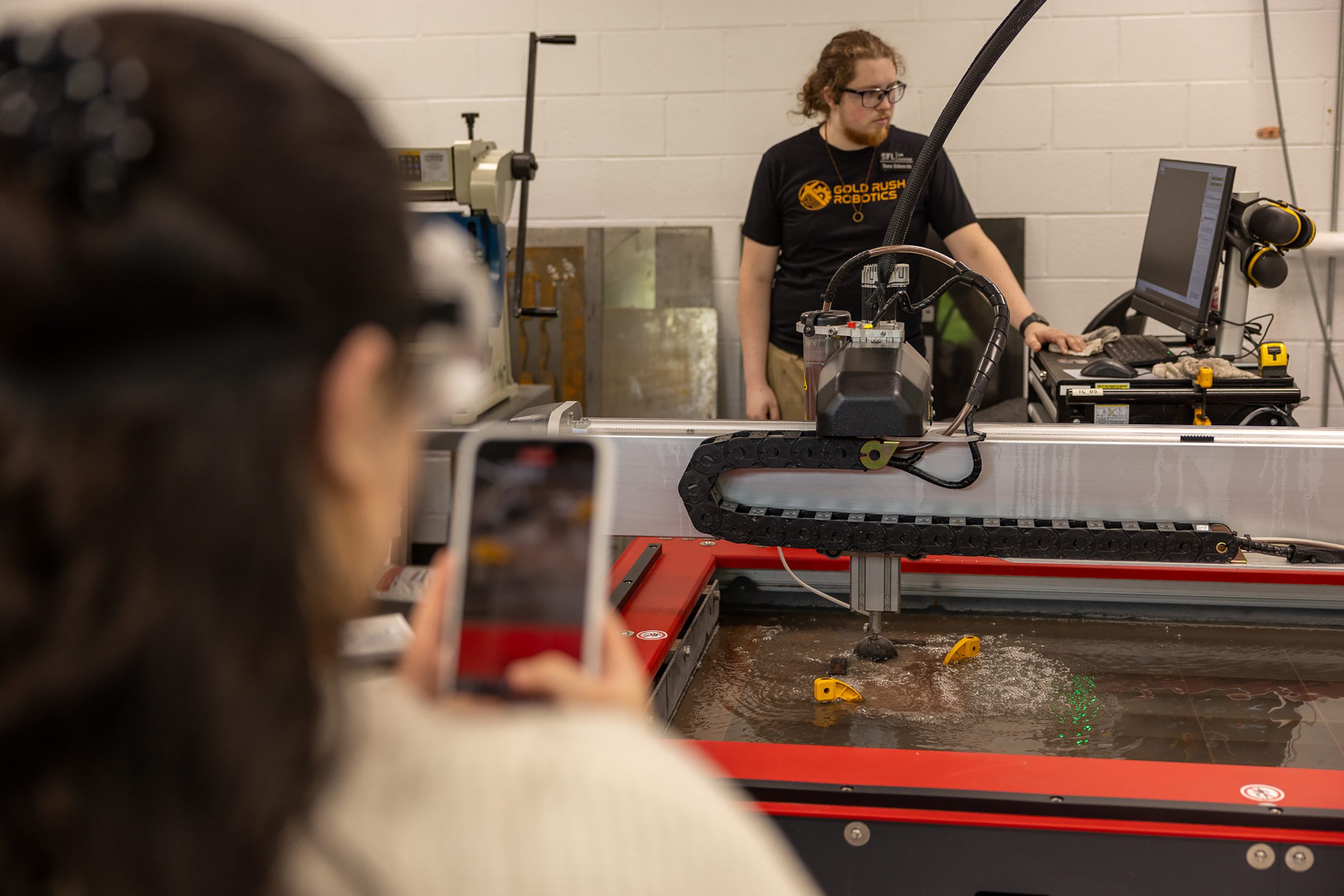 Person uses a phone to record a waterjet cutting machine while a technician operates equipment in a fabrication lab.