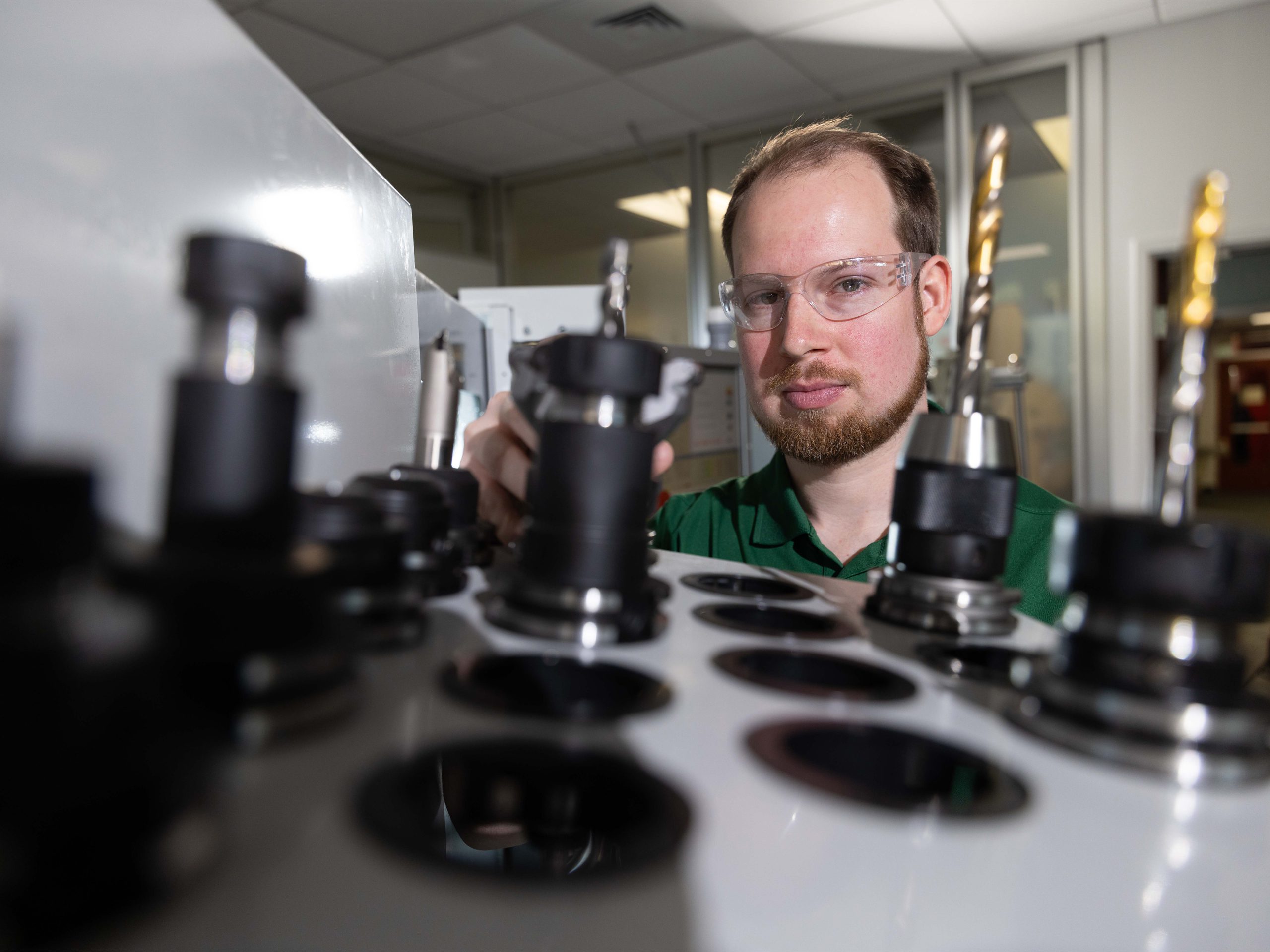 A student wearing safety glasses adjusts a tool holder on a machine, with multiple drill bits and tool slots visible in the foreground of a manufacturing lab.
