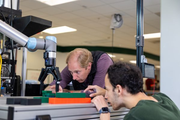 Two people closely inspect and adjust a robotic arm working on a small assembly in a lab setting.