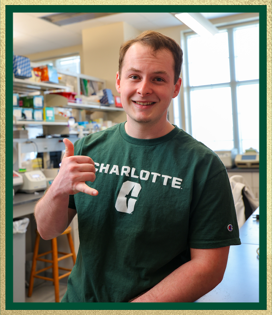 Smiling student in a green Charlotte T-shirt makes a hand gesture while standing in a laboratory with equipment and shelves in the background.