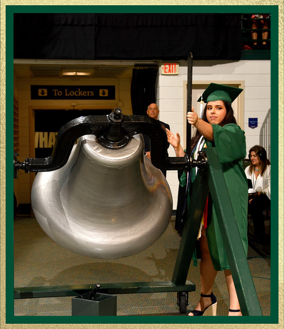 Graduate in a green cap and gown rings a large ceremonial bell indoors during a commencement event, with people and signage visible in the background.