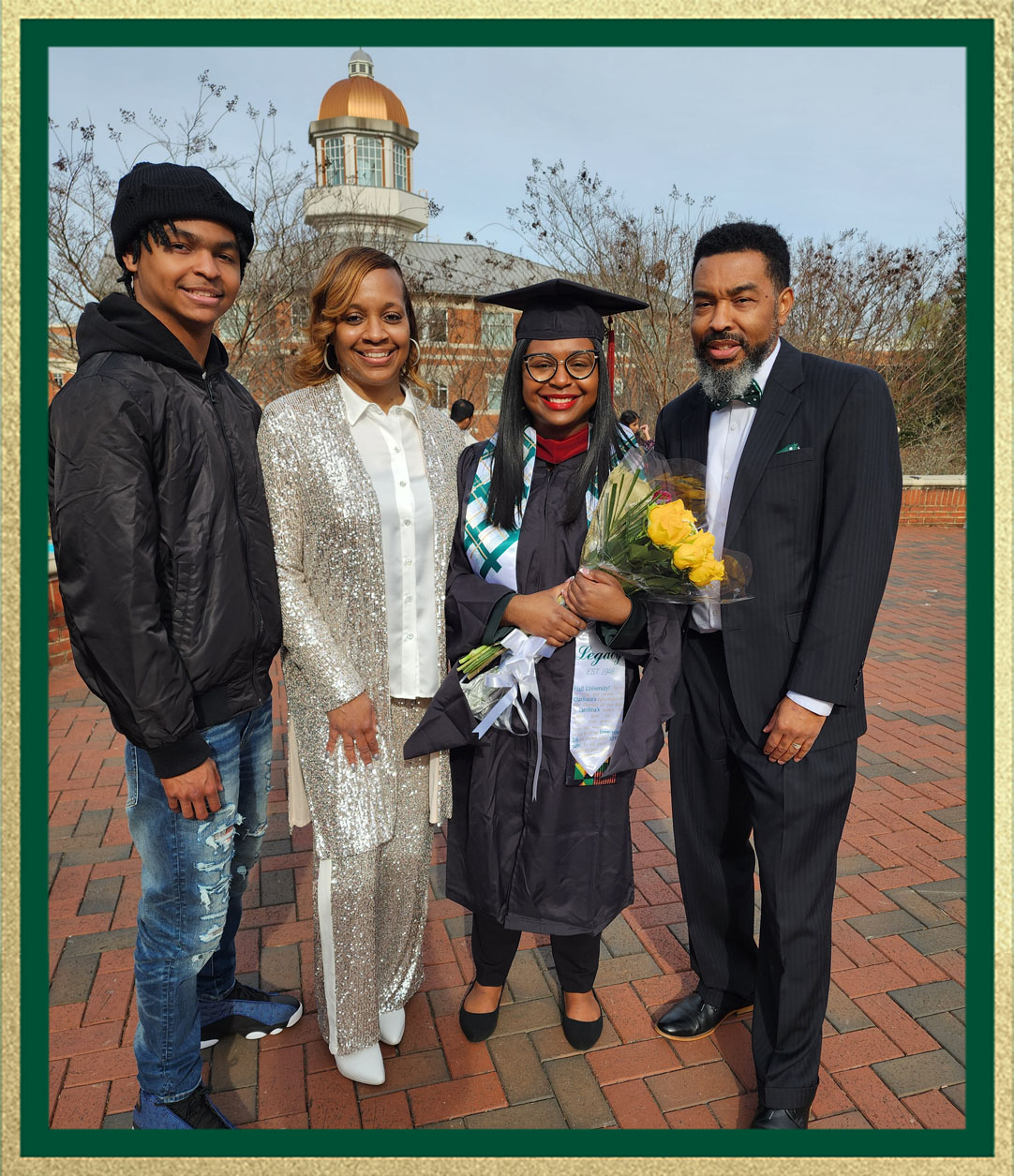 Graduate in cap and gown holds a bouquet of flowers while posing with three family members on a brick campus walkway, with a domed building in the background.