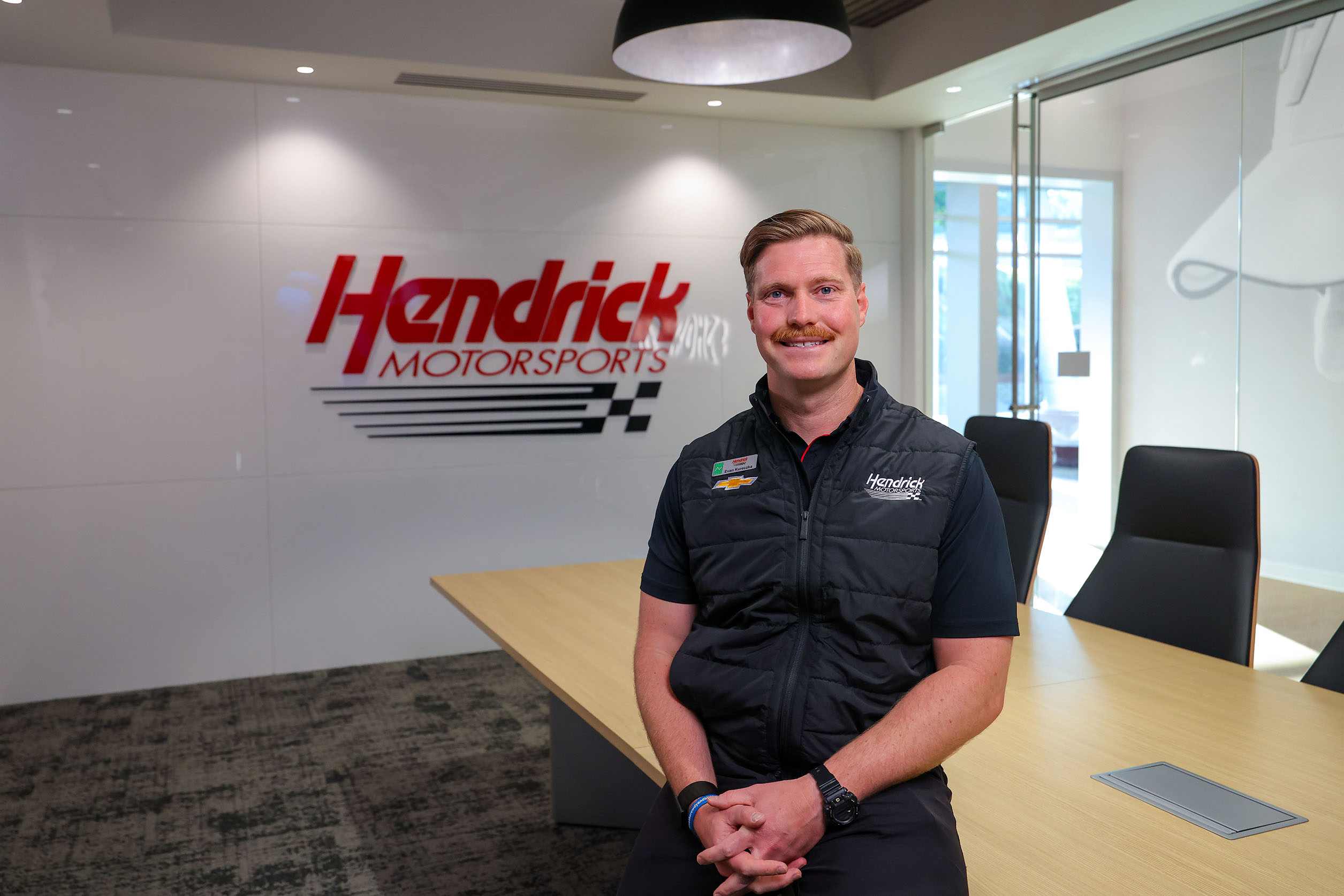 A Hendrick Motorsports performance engineer sits at a conference table in a modern meeting room, smiling in front of a Hendrick Motorsports logo displayed on the wall.