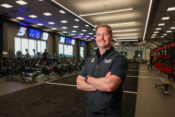 A Hendrick Motorsports staff member stands with arms crossed inside a modern training and performance facility equipped with cardio machines, weightlifting stations, and digital timing displays.
