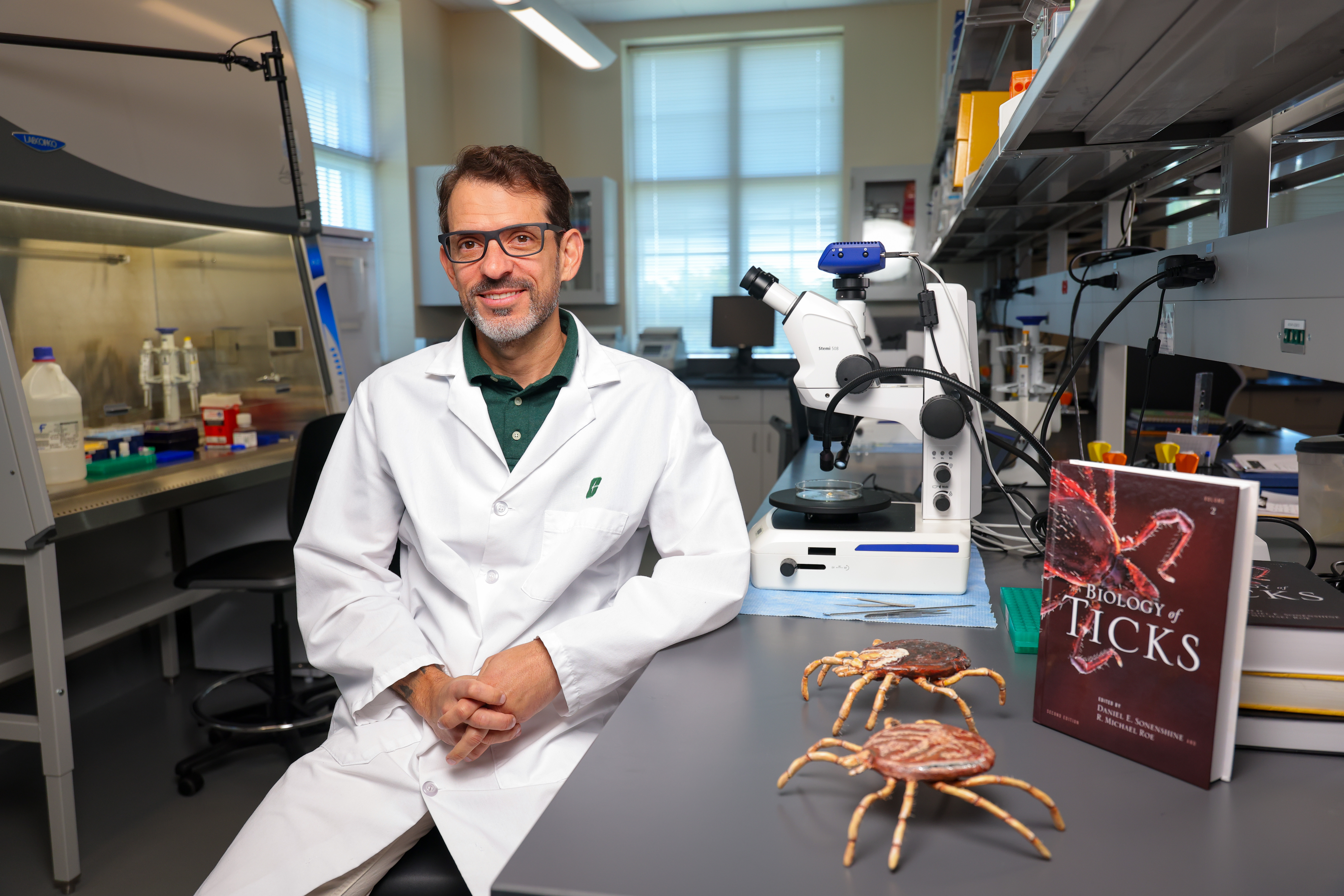 SS_Tick_Hunters_26040_8128_kpl Portrait photo of Rafael Vieira in his lab next to models of ticks and a textbook titled