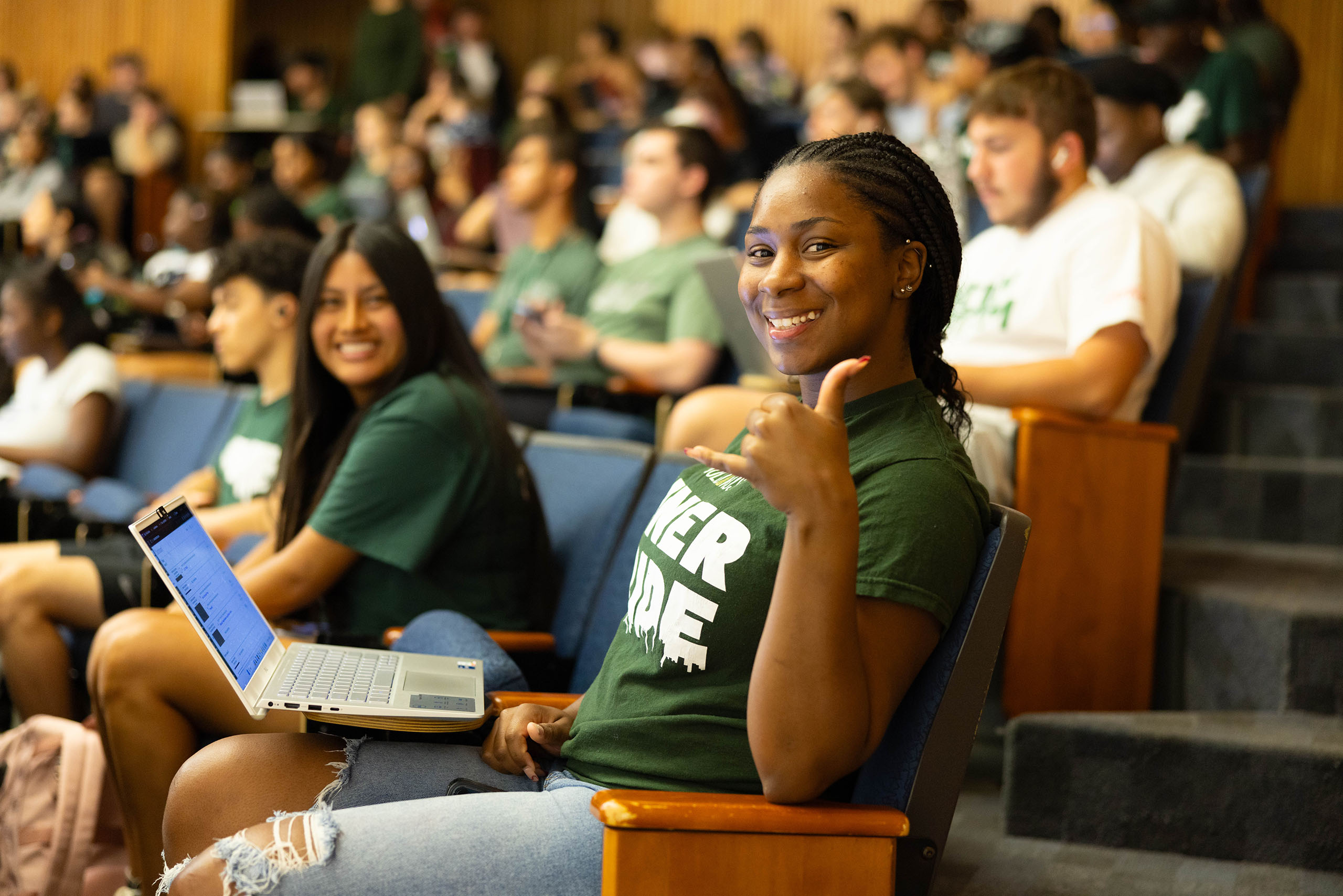 Students-hPeach_Truist-Report A smiling student seated in a lecture hall gives a shaka hand gesture while working on a laptop. Other students fill the classroom in the background, creating an engaged, welcoming campus learning environment.