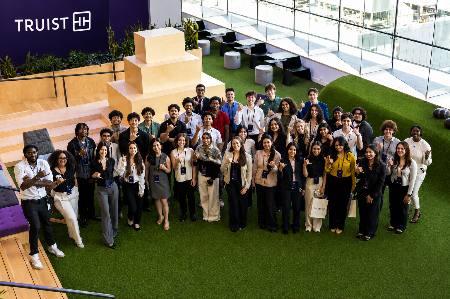 2026 Truist Report - Atrium Large group of students and young professionals pose together inside a Truist office atrium, standing on a green turf area beneath a Truist sign and giving thumbs-up gestures.