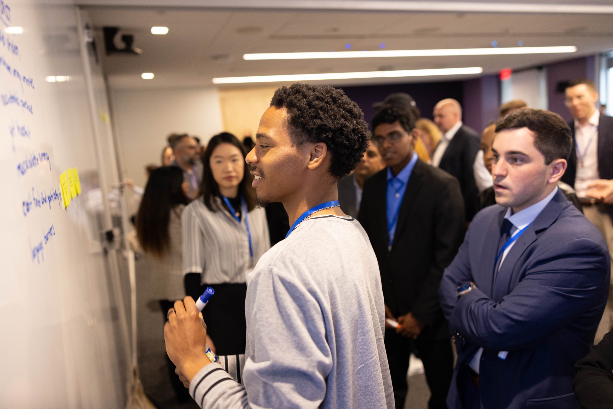 2026 Truist Report - Immersive 2 Participant writes ideas on a whiteboard during a collaborative workshop while a group of students and professionals gather around to discuss and review the brainstorming session.