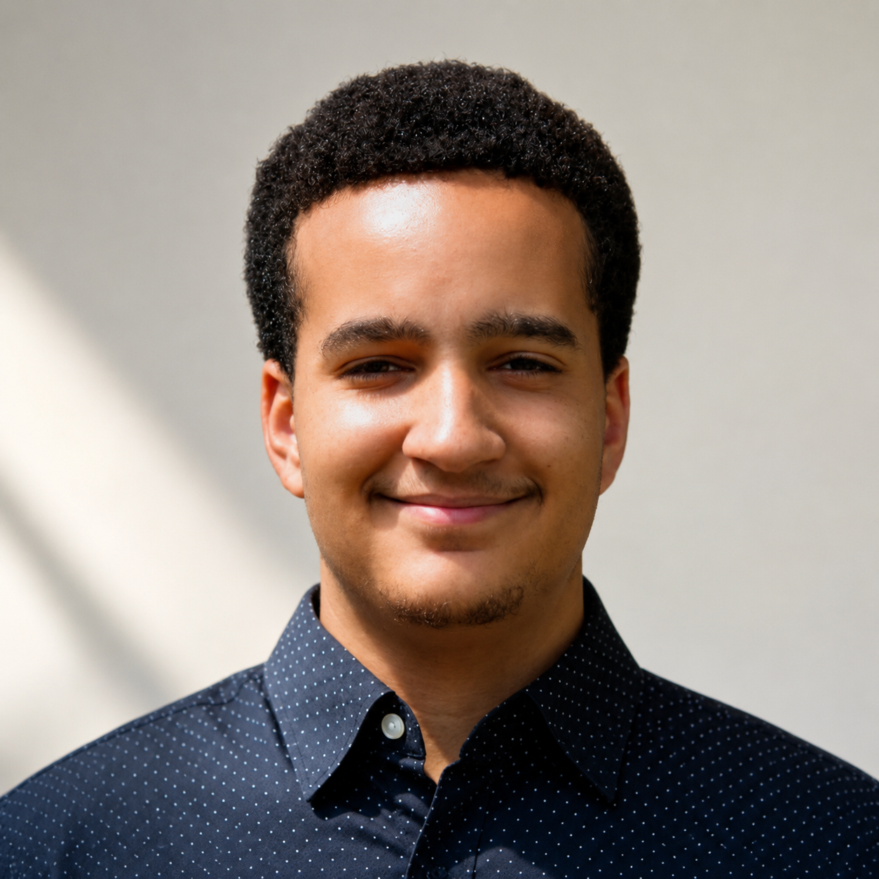 A person with short curly hair smiles at the camera while wearing a dark blue button-down shirt with a subtle dotted pattern against a softly lit background.