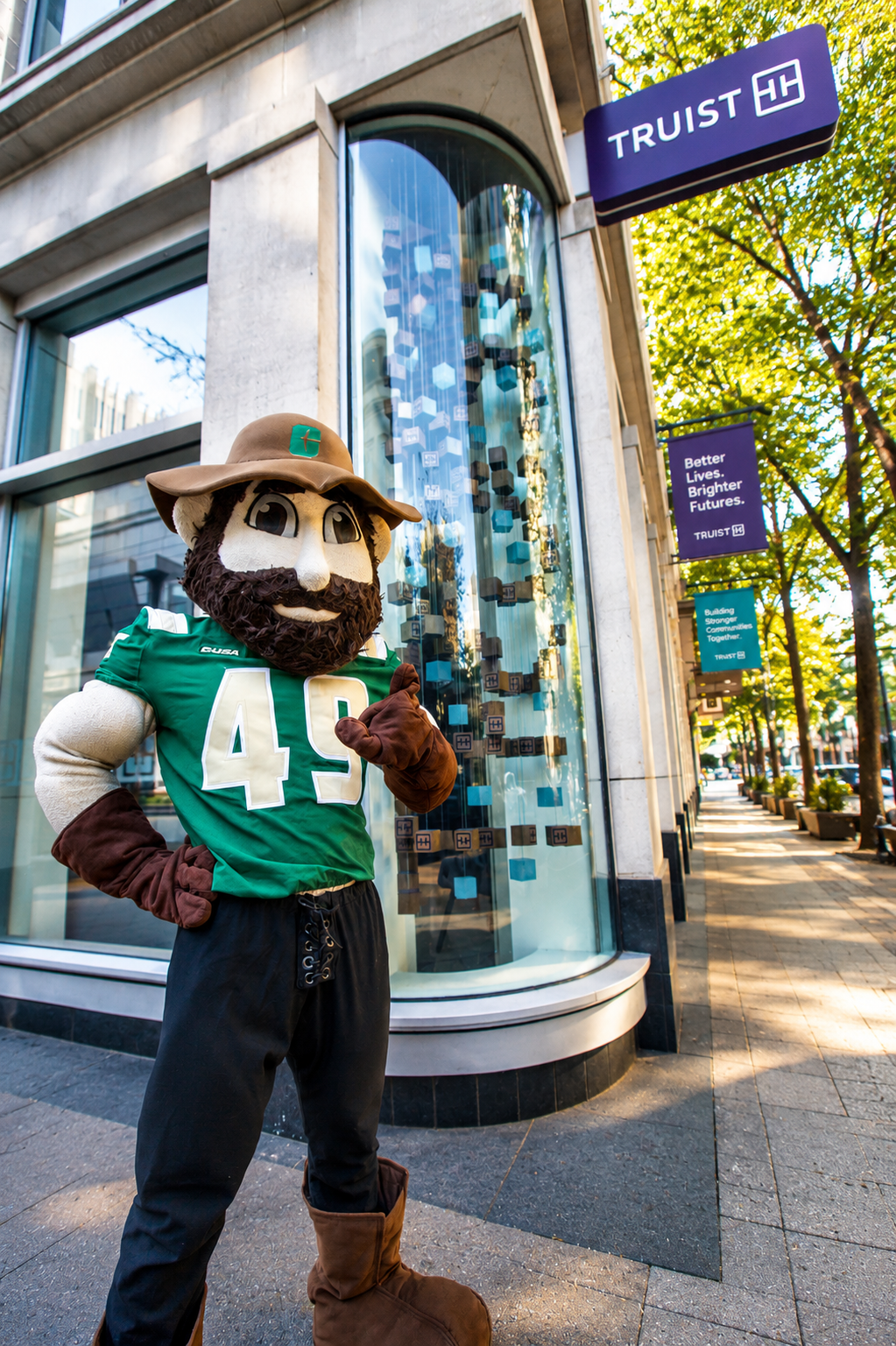 2026 Truist Report - Norm UNC Charlotte’s mascot, Norm the Niner, stands outside a Truist bank branch on a city sidewalk, posing in a green jersey beneath Truist signage.