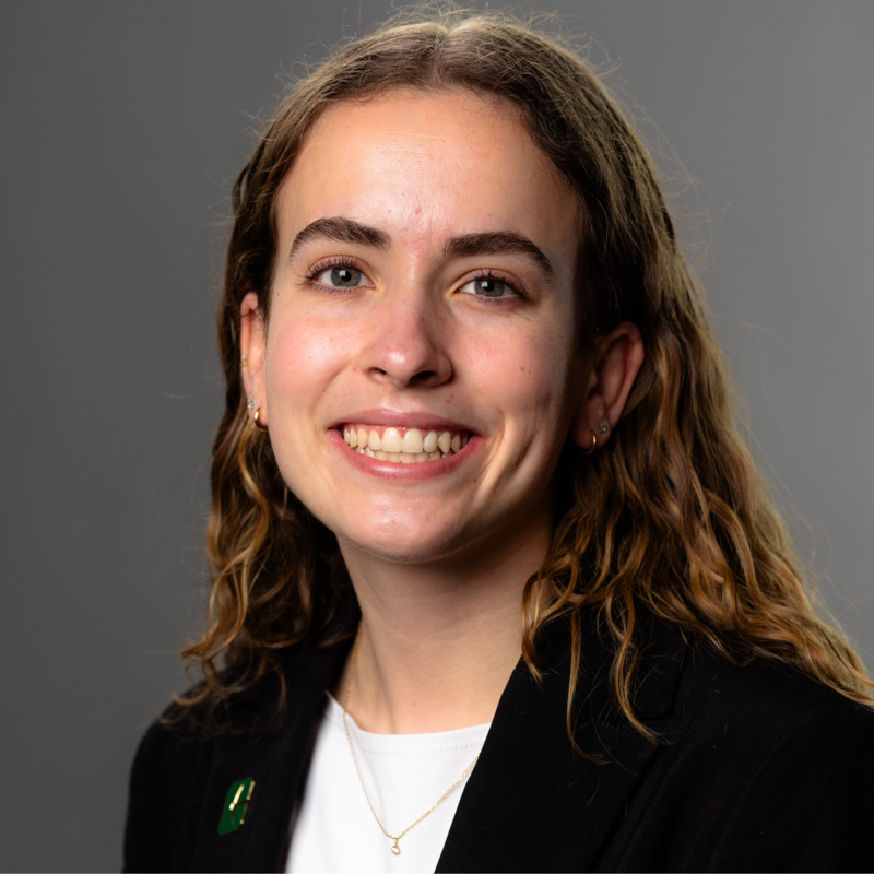 A person smiles in a professional headshot while wearing a black blazer over a white top with a green lapel pin against a gray studio background.